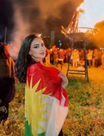 Draped in the Kurdish flag, a young Kurdish woman stands before the Newroz fire in Basur (Kurdistan in Iraq). Photo: Instagram/SA
